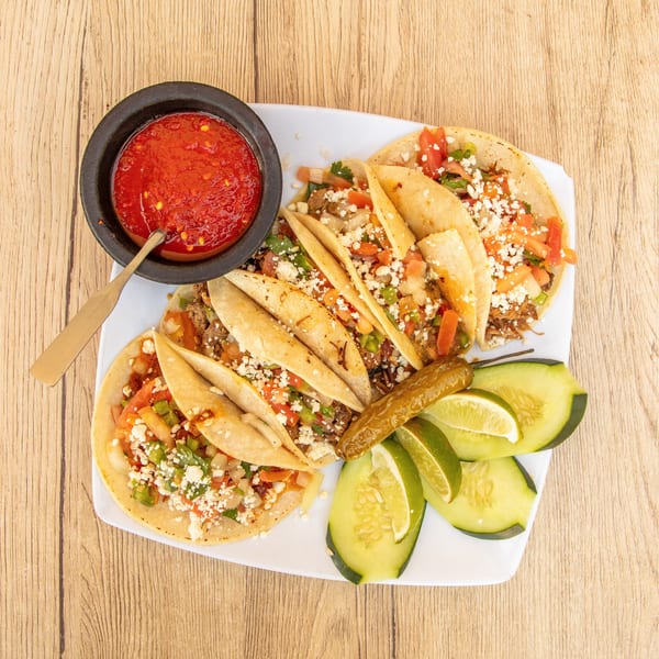 A plate of tacos with salsa, cucumber slices, lime wedges, and a pickled pepper on a wooden table.