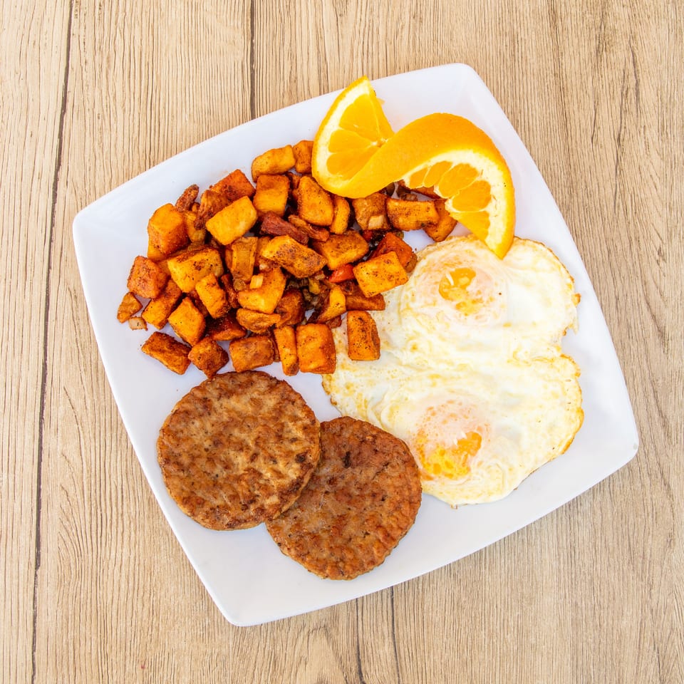 Breakfast plate with two fried eggs, two sausage patties, seasoned potato cubes, and orange slices.