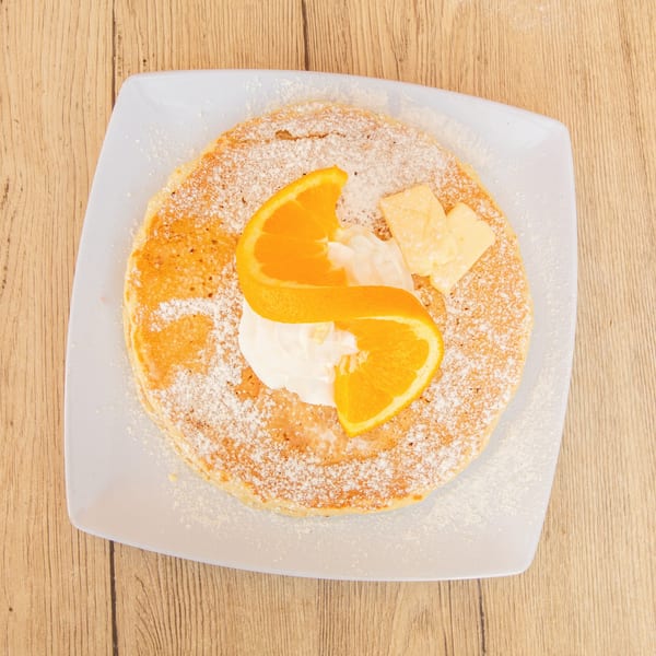 Overhead view of a pancake with powdered sugar, whipped cream, orange slices, and butter.