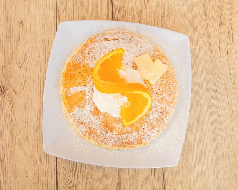 Overhead view of a pancake with powdered sugar, whipped cream, orange slices, and butter.