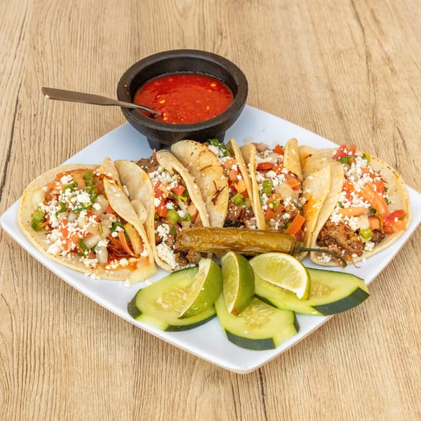 A plate of six tacos with meat, cheese, and salsa, served with limes, cucumbers, and a side of red salsa.