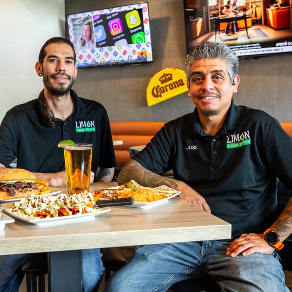 Two men in Limón Urban Kitchen shirts smile at a table laden with food and drinks.