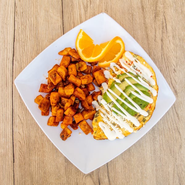 A breakfast plate with a fluffy omelet topped with avocado and sour cream, alongside seasoned potato cubes and orange slices.