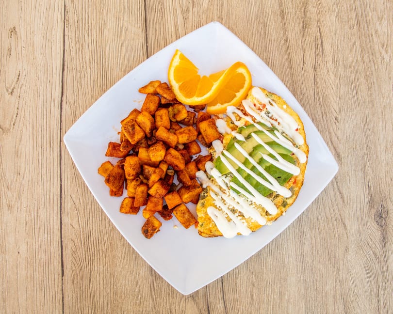 A breakfast plate with a fluffy omelet topped with avocado and sour cream, alongside seasoned potato cubes and orange slices.