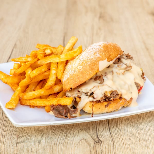 Cheesesteak sandwich with melted cheese and fries on a white plate, on a wooden table.