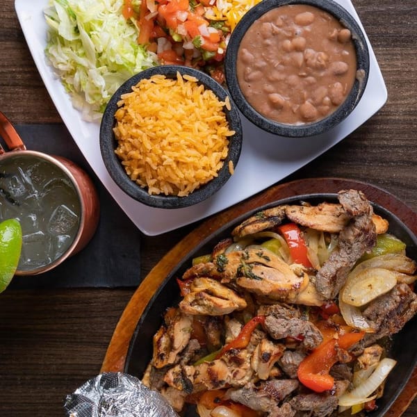 Sizzling fajitas with chicken and steak, rice, beans, and a margarita on a wooden table.