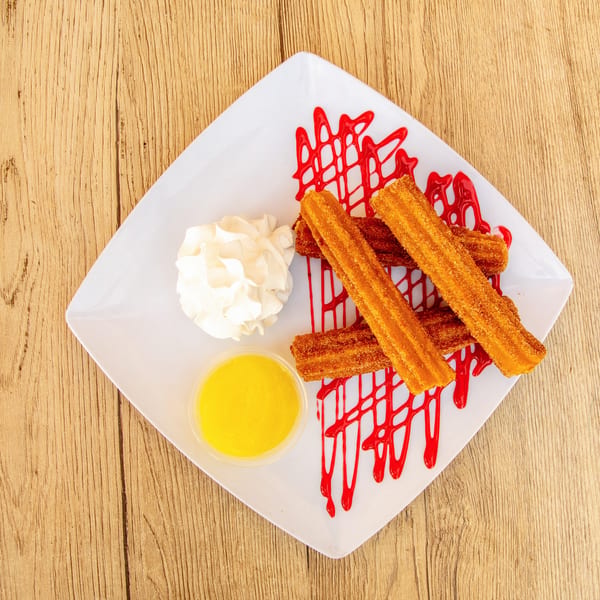 Churros with red drizzle, whipped cream, and a side of yellow sauce on a white plate.