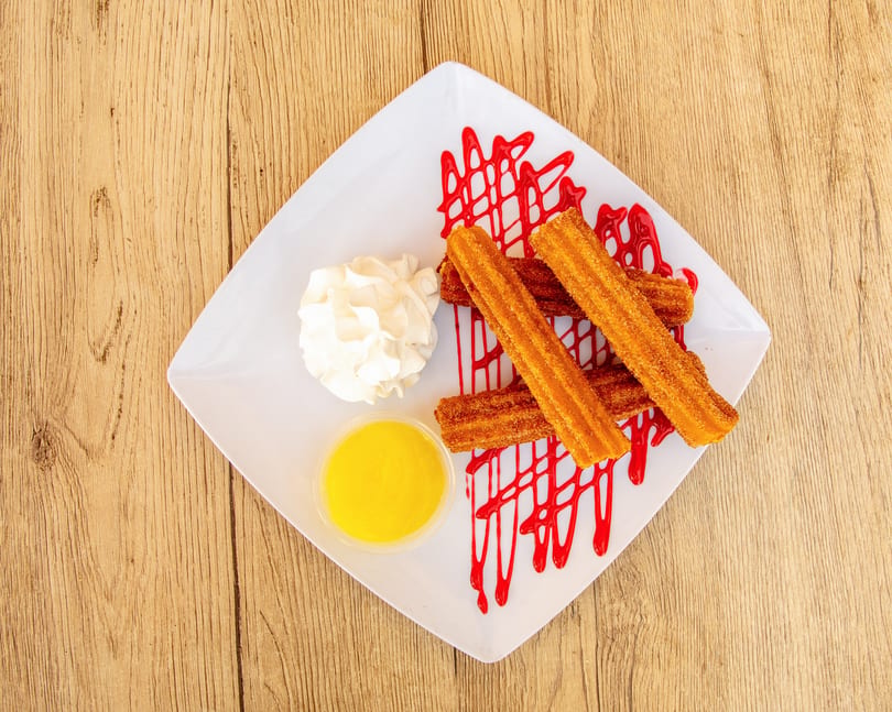 Churros with red drizzle, whipped cream, and a side of yellow sauce on a white plate.